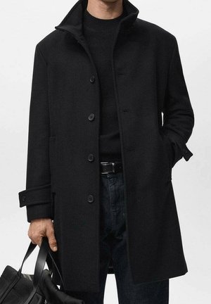 Mann trägt schwarzen, geknöpften Mantel und Pullover, dunkle Jeans, hält eine schwarze Tasche in einer Hand, andere Hand in der Manteltasche.