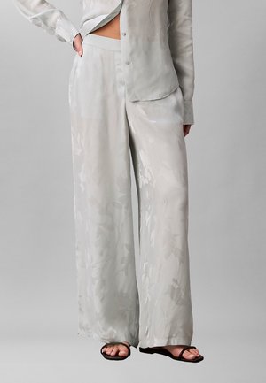 Femme portant un pantalon large en satin gris clair et une chemise assortie partiellement déboutonnée, avec des sandales noires à brides sur fond gris uni.