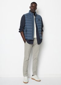 Wattierte marineblaue Weste über einem dunklen Hemd, kombiniert mit hellbeigen Hosen und weißen Sneakers mit braunen Akzenten. Mit Reißverschluss ausgestattete Taschen auf der Weste.