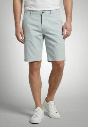 Man wearing light gray knee-length shorts, white sneakers, and a white shirt against a plain gray background.
