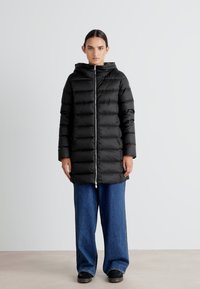 Zwarte puffercoat met capuchon, voorzien van een ritssluiting aan de voorkant, horizontale stiksels en een knielange ontwerp. Draagt over wijde denim.