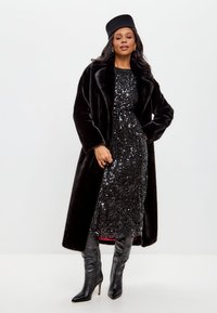 Zwarte nepbontjas met brede revers, gedragen over een zwart paillettenjurk. Knielange zwarte laarzen met hakken en een bijpassende zwarte hoed maken de look compleet.