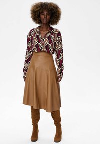 Woman with curly hair wearing a patterned long-sleeve blouse, tan leather midi skirt, and knee-high suede brown boots standing against white background.