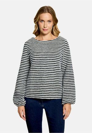 Femme portant un pull à manches longues rayé horizontalement noir et blanc avec des manches bouffantes et un jean bleu foncé, debout devant un fond blanc.