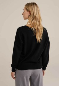Maglione nero lavorato a maglia con una vestibilità morbida, spalle basse, polsini e orlo a costine. Tessuto morbido, design minimalista, indossato con pantaloni grigi.