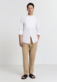 Mann steht da und trägt ein weißes Hemd mit Mandarin-Kragen und hochgekrempelten Ärmeln, beige strukturierte Hose und dunkle gewebte Sandalen vor einem weißen Hintergrund.
