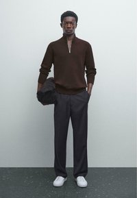 Dunkelbrauner, gerippter Strickpullover mit halbem Reißverschluss-Kragen, kombiniert mit grauen, maßgeschneiderten Hosen und weißen Sneakers, gehalten mit einer schwarzen Tasche.
