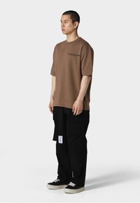 Brun oversized t-shirt med korte ærmer og sort logo, parret med sorte cargo bukser med lommer og et mærke. Sorte sneakers fuldender looket.