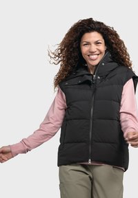 Schwarze, gesteppte Weste mit hohem Kragen und frontalem Reißverschluss, getragen über einem rosa Langarmshirt, kombiniert mit hellen Hosen.