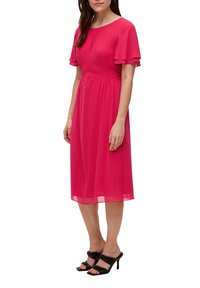 Rosa Chiffonkleid mit kurzen, schwingenden Ärmeln, gerafftem Taillenbereich und knielangem Saum. Kombiniert mit schwarzen, schmalen, hohen Sandalen.