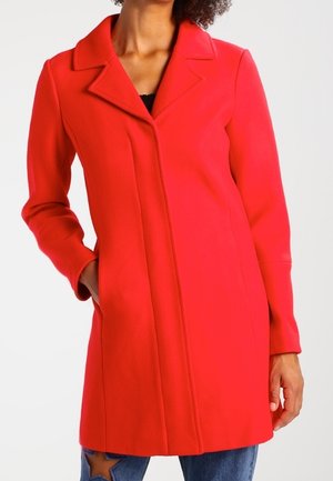 Vrouw draagt een felrode, knielange jas met een brede kraag en zijzakken, gecombineerd met een blauwe spijkerbroek met een sterpatch.