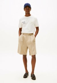 Camisa de algodón blanca con logotipo bordado, pantalones cortos de rayas beige con plisados y mocasines marrón oscuro. El modelo lleva una gorra azul.