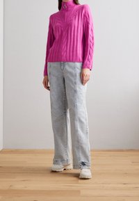 Pull en maille torsadée rose avec col montant et fermeture éclair argentée, associé à un jean ample délavé clair et des baskets blanches sur un sol en bois.