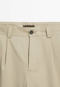 Pantaloni beige con fronte piatto, dotati di due tasche laterali, chiusura con un bottone e passanti per cintura. Realizzati in tessuto leggero e liscio.