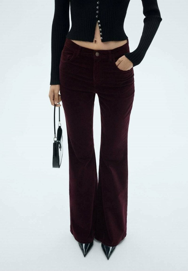 Femme portant un pantalon flare en velours côtelé bordeaux foncé, un pull court noir boutonné, des chaussures noires pointues, tenant un petit sac à main noir.
