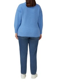 Kobieta ubrana w jasnoniebieski sweter, niebieskie dżinsy i białe buty, stojąca tyłem na tle jednolitej białej ściany.