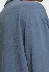 Chemise bleue texturée à coupe décontractée, fabriquée dans un tissu doux froissé, avec un col classique. Détails comprenant des coutures et des surpiqûres visibles.