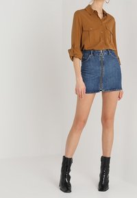 Donna che indossa una camicia marrone con bottoni, maniche arrotolate, una minigonna in denim blu con zip frontale e stivali alla caviglia in patent leather neri.