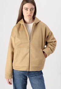 Beige zip-up jakke laget av myk fleece, med høy krage, sidelommer og avslappet passform. Brukt over en lys topp.