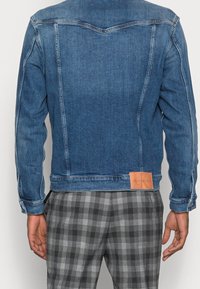 Chaqueta de mezclilla azul oscura con un diseño de costura en la parte posterior, con una etiqueta de cuero en el dobladillo. Combinada con pantalones de cuadros grises.