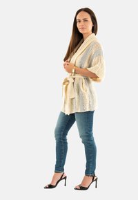 Cardigan a righe beige e grigio in maglia con cintura, dotato di maniche corte e apertura frontale. Indossato sopra jeans blu e tacchi neri.