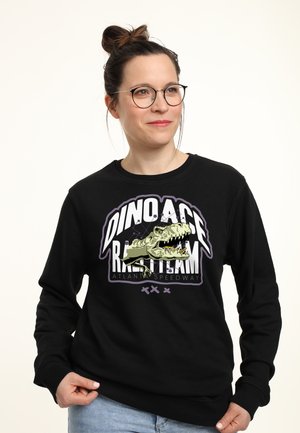 Kobieta w okularach, która ma na sobie czarną bluzę z grafiką dinozaura i napisem „Dino Ace R.A. Team Atlanta Speedway”, uśmiechająca się i patrząca w prawo.