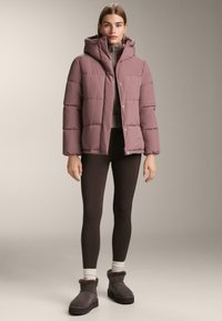 Kurtka puchowa w delikatnym kolorze mauve, wyposażona w kaptur, pikowany wzór i zapinana na zatrzaski. Połączona z ciemnymi legginsami i kozakami z futrzanym obszyciem.