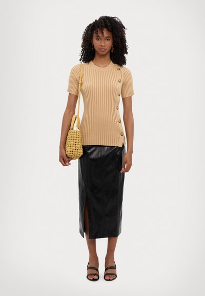 Beige gerippter Stricktop mit Knöpfen, kombiniert mit einem schwarzen Midirock aus Kunstleder mit seitlichem Schlitz. Das Model hält eine gelbe geflochtene Tasche.