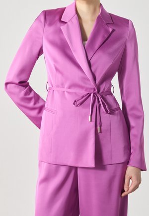 Femme portant un blazer rose vif cintré avec une ceinture nouée et un pantalon rose assorti, debout devant un fond uni.