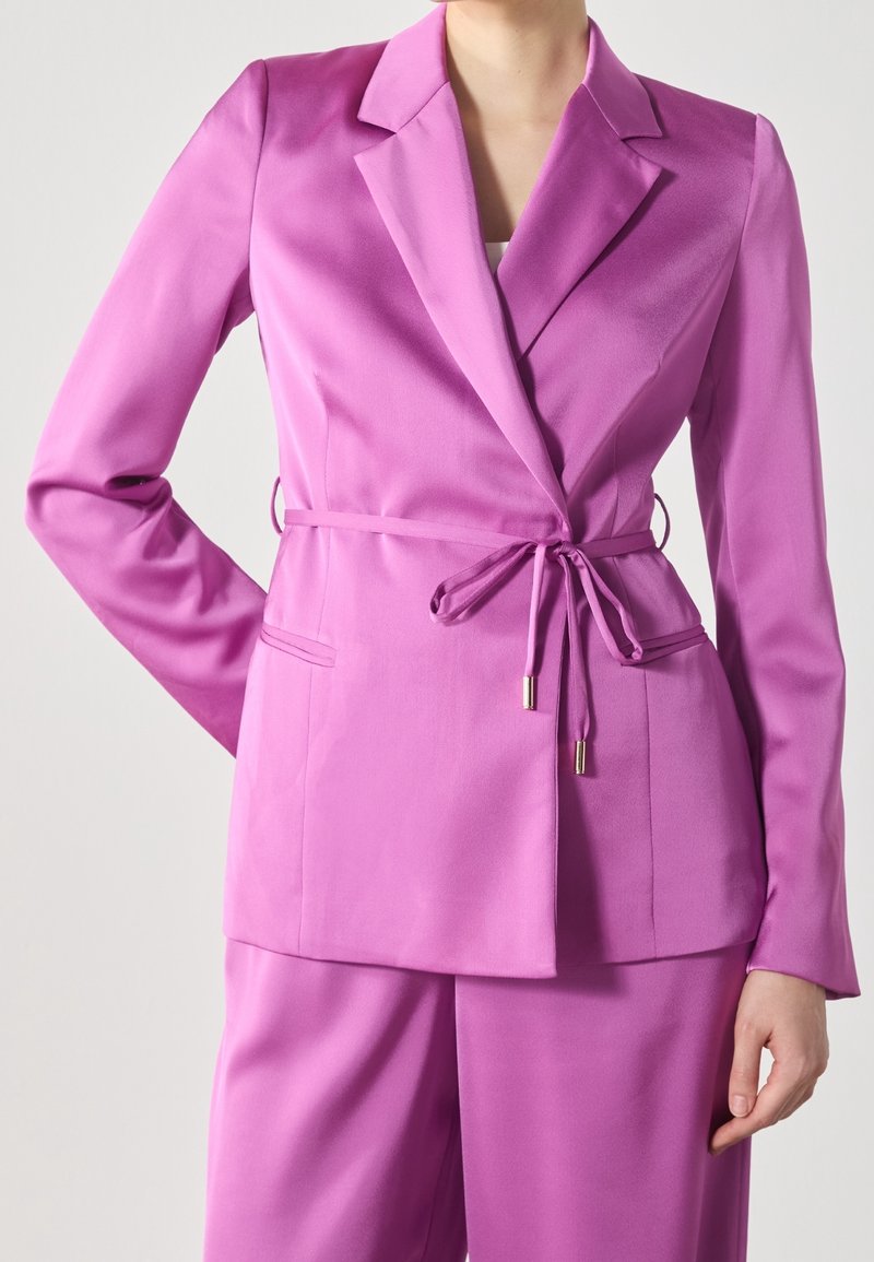Femme portant un blazer rose vif cintré avec une ceinture nouée et un pantalon rose assorti, debout devant un fond uni.