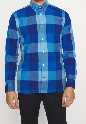 Homme portant une chemise à grands carreaux bleus et marine à manches longues et un pantalon noir, debout devant un fond blanc.