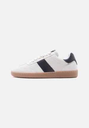 Baltas zomšinis sportinis batelis su rudos gumos padais ir juoda odine juostele bei kulno kilpele, vaizduojamas šone ant balto fono.