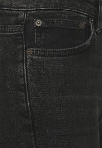 Jean en denim noir avec une texture subtile, cinq poches et une quincaillerie métallique distinctive à finition polie.