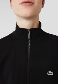 Primo piano di una persona che indossa una giacca nera con zip e colletto alto, con il logo del coccodrillo Lacoste sul lato sinistro del petto.