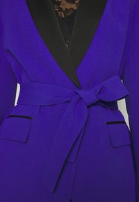 Blazer violet avec revers noirs, ceinture à nouer à la taille, deux poches à rabat à l'avant, porté sur un haut en dentelle noire.