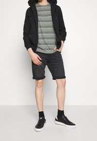 Czarna bluza z kapturem na zamek, wzorzysta zielona koszulka, czarne krótkie dżinsy i czarne trampki z białą podeszwą, stojący na neutralnym tle.