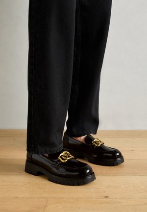 Personne portant des mocassins en cuir verni noir avec semelles épaisses et détail chaîne dorée, assortis à un pantalon droit noir sur un sol en bois.