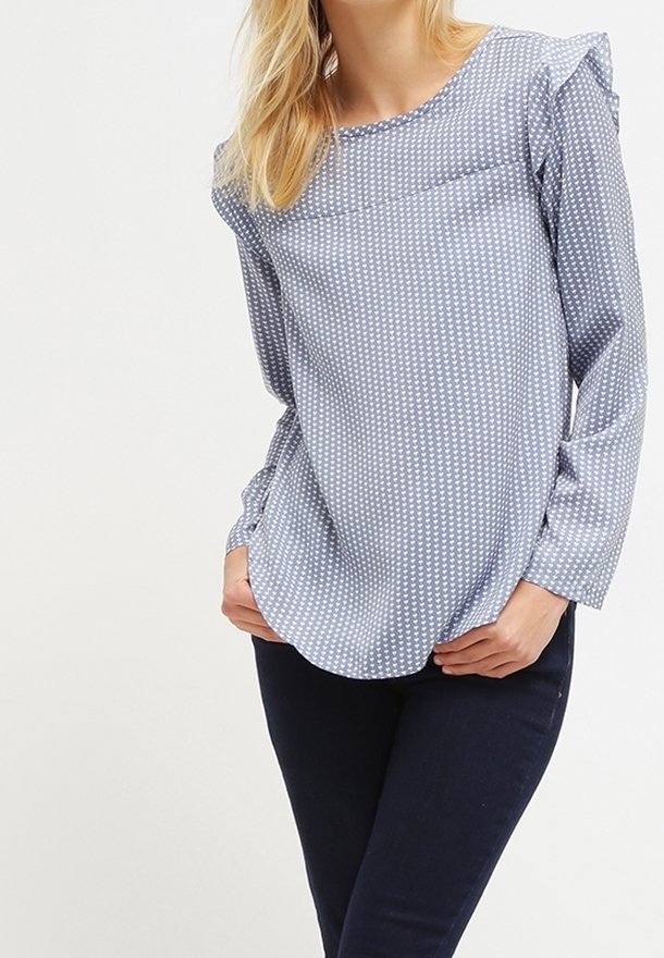 Femme portant un chemisier bleu clair à manches longues avec de petits motifs blancs, associé à un pantalon bleu foncé, les mains reposant près des poches.