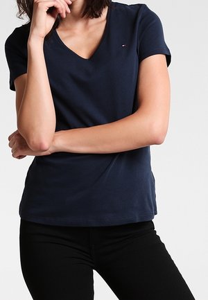 Kobieta ubrana w granatowy T-shirt z krótkim rękawem i dekoltem w serek z małym logo na piersi, z założonymi rękami, w połączeniu z czarnymi spodniami.