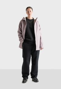 Giacca impermeabile viola chiaro con cappuccio sopra un maglione nero, abbinata a pantaloni neri e scarpe nere.