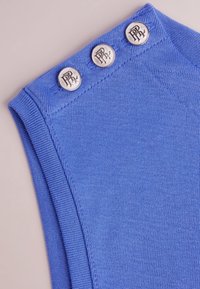 Blå stickad tyg med en slät textur har tre metallknappar med ingraverade logodetaljer längs axelsömmen.