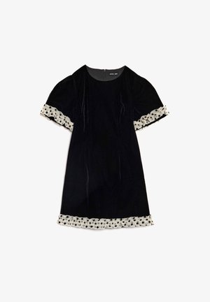 Juoda aksominė suknelė su trumpomis pūstomis rankovėmis, kurių rankogaliuose ir suknelės apačioje yra baltų polka taškuotų nėrinių apvadai, užtrauktuku nugarinėje dalyje.