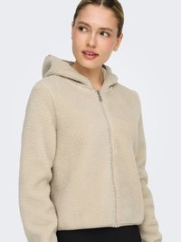 Woman wearing a beige sherpa fleece zip-up hoodie with a hood, looking to her left against a plain white background.