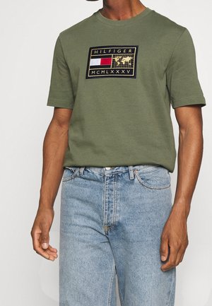 T-shirt vert olive à manches courtes, avec un logo brodé rectangulaire en rouge, blanc et bleu marine. Porté avec un jean en denim bleu clair.