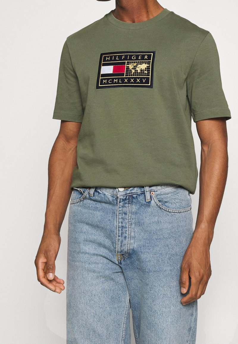 Camiseta verde oliva de manga corta, con un logotipo bordado rectangular en rojo, blanco y azul marino. Combinada con jeans de mezclilla azul claro.