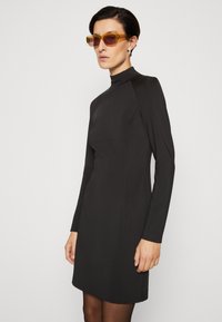 Robe noire à manches longues avec un col montant, une silhouette ajustée et un tissu lisse. Portée avec des lunettes de soleil jaunes et des collants transparents.