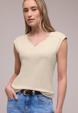 Femme aux longs cheveux blonds portant un haut beige sans manches et un jean bleu avec une ceinture marron, debout devant un fond uni.