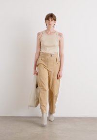 Beige top tank ja löysähköt vaaleanruskeat housut, joissa on sivutaskut. Malli kantaa valkoista käsilaukkua ja käyttää vaaleanharmaita nilkkureita.