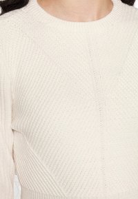 Krémový pletený svetr s žebrovanou texturou, kulatým výstřihem a detailní diagonální výšivkou. Tkanina je měkká a zajišťuje těsný fit.