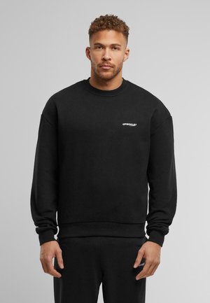 Schwarzer Sweatshirt aus weichem Stoff mit Rundhalsausschnitt, langen Ärmeln und einem kleinen weißen Logo auf der Brust. Schlichtes Design und Passform.